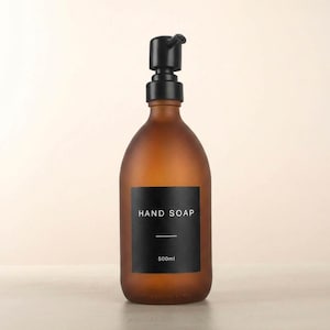 Może przedstawiać: Brązowy szklany dozownik mydła do rąk z czarną pompką. Dozownik ma czarną etykietę z napisem "HAND SOAP" i "500ml". Butelka ma matowe wykończenie i zaokrąglony kształt.