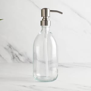 May include: Clear glass soap dispenser with a brushed metal pump. The bottle has a rounded shape and a long, white plastic tube. The dispenser is empty and sits on a white marble surface.