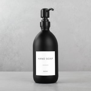 May include: A matte black hand soap dispenser with a pump top. A white rectangular label on the front reads "HAND SOAP" with a line underneath and "500ml" below. The bottle is on a light gray surface.