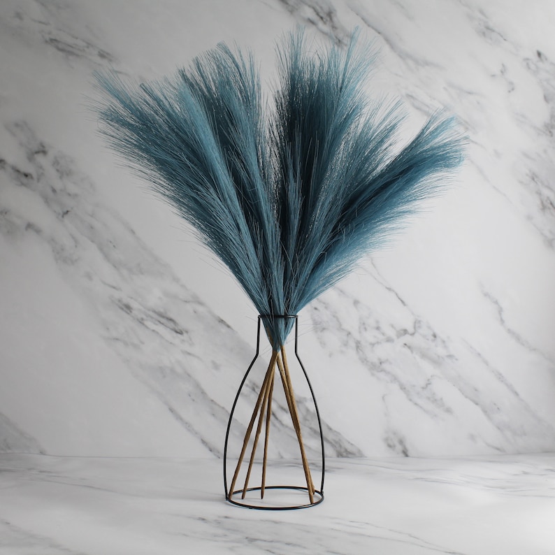 May include: A bouquet of teal-colored faux pampas grass in a black metal vase with gold-toned stems. The vase has a minimalist, open design. The background is a white marble pattern.