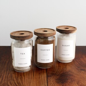K&ouml;nnte beinhalten: Drei Glasbeh&auml;lter mit Holzdeckeln und Etiketten. Die Beh&auml;lter sind mit Tee, Kaffee und Zucker gef&uuml;llt. Die Etiketten lauten "Tee", "Kaffee" und "Zucker" mit dem Namen "NAMIE HOME" unter jedem Etikett.
