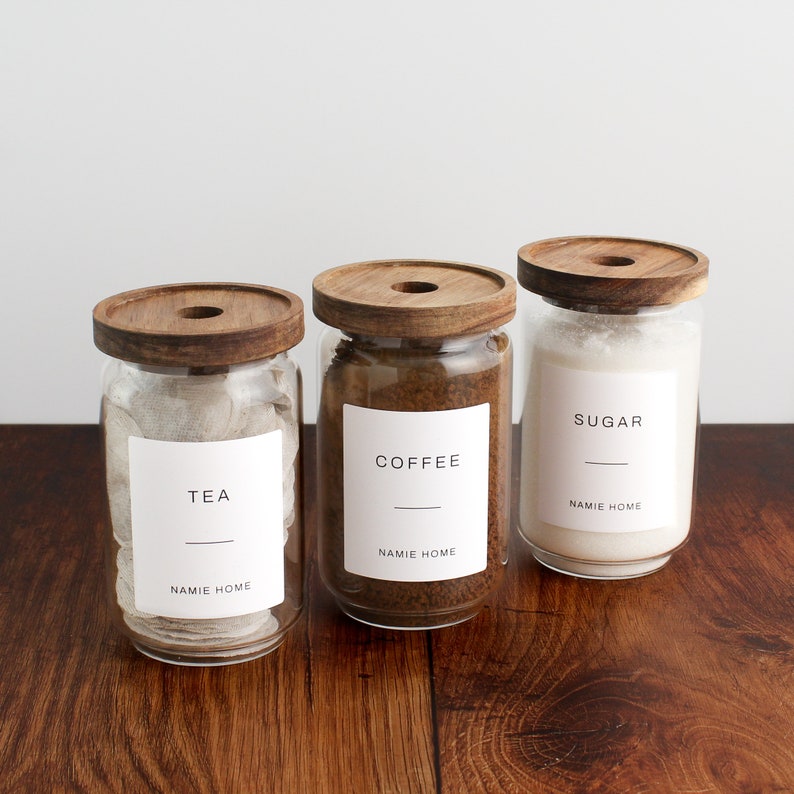 K&ouml;nnte beinhalten: Drei Glasbeh&auml;lter mit Holzdeckeln und Etiketten. Die Beh&auml;lter sind mit Tee, Kaffee und Zucker gef&uuml;llt. Die Etiketten lauten "Tee", "Kaffee" und "Zucker" mit dem Namen "NAMIE HOME" unter jedem Etikett.