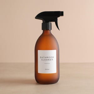 Pode incluir: Um borrifador de vidro castanho com um gatilho preto. A garrafa tem um rótulo branco que diz "BATHROOM CLEANER" e "500ml". A garrafa foi projetada para limpar banheiros.
