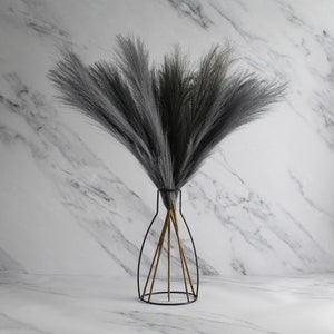 May include: A decorative arrangement of gray faux pampas grass in a black metal vase with a geometric design. The vase has a slender neck and a wide base, with wooden supports. The background is a white marble pattern.