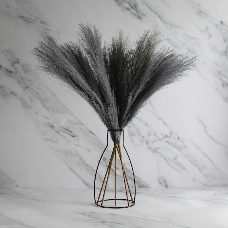 May include: A decorative arrangement of gray faux pampas grass in a black metal vase with a geometric design. The vase has a slender neck and a wide base, with wooden supports. The background is a white marble pattern.