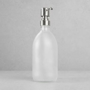 Puede incluir: Un dispensador de jabón de vidrio esmerilado con una bomba de acero inoxidable. La botella tiene una forma redondeada y un acabado mate. La bomba tiene una boquilla curva para dispensar jabón líquido.