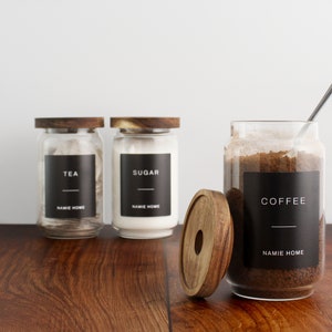 Puede incluir: Tres tarros de vidrio con tapas de madera y etiquetas negras. Las etiquetas dicen "TEA", "SUGAR" y "COFFEE". Cada etiqueta tambi&eacute;n incluye el texto "NAMIE HOME".