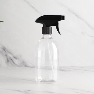 Puede incluir: Un pulverizador de plástico transparente con gatillo y boquilla negros. La botella tiene forma redondeada y un tubo largo y fino en el interior. El fondo es una superficie de mármol blanco.