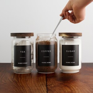 Puede incluir: Tres tarros de vidrio con tapas de madera y etiquetas negras. Las etiquetas dicen "T&eacute;", "Caf&eacute;" y "Az&uacute;car". Los tarros est&aacute;n llenos de bolsitas de t&eacute;, granos de caf&eacute; y az&uacute;car.