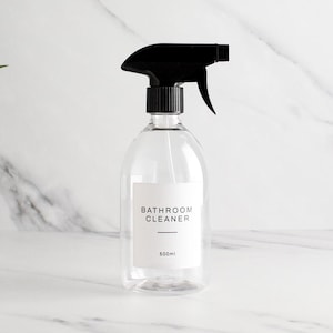 以下が含まれることがあります： 黒いトリガーが付いた透明なプラスチック製のスプレーボトル。白いラベルには「BATHROOM CLEANER」と「500ml」と書かれています。 ボトルは掃除用に設計されており、白い大理石の背景に配置されています。