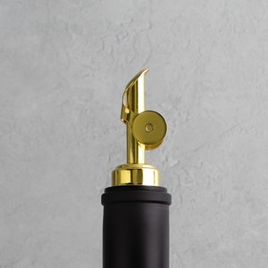 May include: A gold bottle stopper with a black top. The stopper has a ring around the top and a spout for pouring.