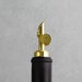 May include: A gold bottle stopper with a black top. The stopper has a ring around the top and a spout for pouring.