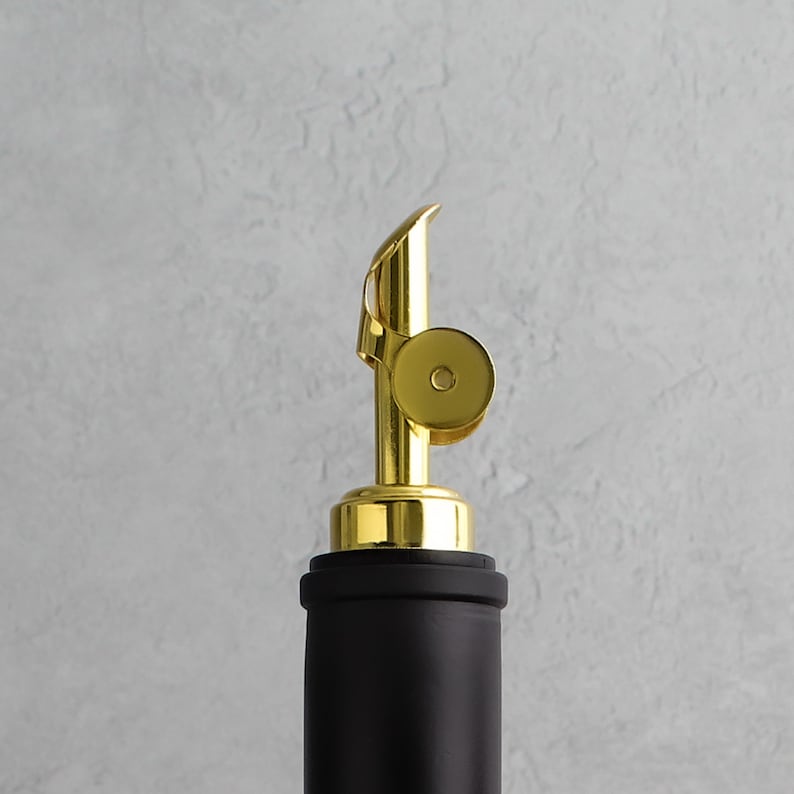 May include: A gold bottle stopper with a black top. The stopper has a ring around the top and a spout for pouring.