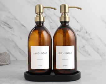 May include: Two amber glass soap dispensers with gold pumps and black labels. The labels read "HAND SOAP 500ml" and "DISH SOAP 500ml". The dispensers are sitting on a black oval tray.