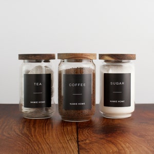 Puede incluir: Tres tarros de vidrio con tapas de madera y etiquetas negras. Las etiquetas dicen "T&eacute;", "Caf&eacute;" y "Az&uacute;car" con el texto "NAMIE HOME" debajo de cada etiqueta.