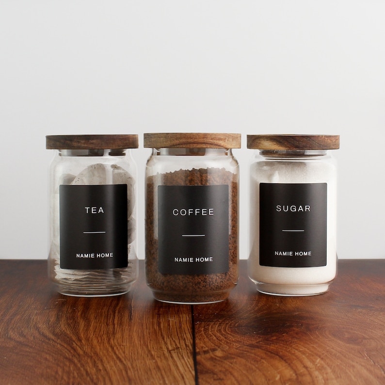 Puede incluir: Tres tarros de vidrio con tapas de madera y etiquetas negras. Las etiquetas dicen "T&eacute;", "Caf&eacute;" y "Az&uacute;car" con el texto "NAMIE HOME" debajo de cada etiqueta.