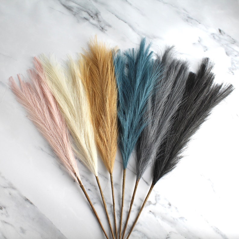May include: A collection of faux pampas grass stems in various colors, including pink, cream, tan, teal, gray, and black. The stems have a feathery texture and are arranged against a white marble background. These decorative stems are suitable for home decor.