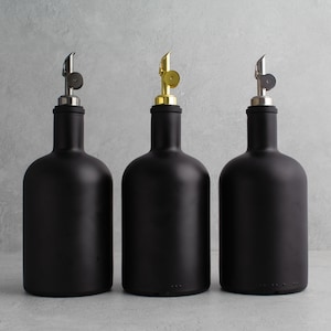 May include: Three black glass bottles with different colored metal tops. The bottles are all the same size and shape, and they are arranged in a row on a gray background.