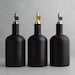 May include: Three black glass bottles with different colored metal tops. The bottles are all the same size and shape, and they are arranged in a row on a gray background.