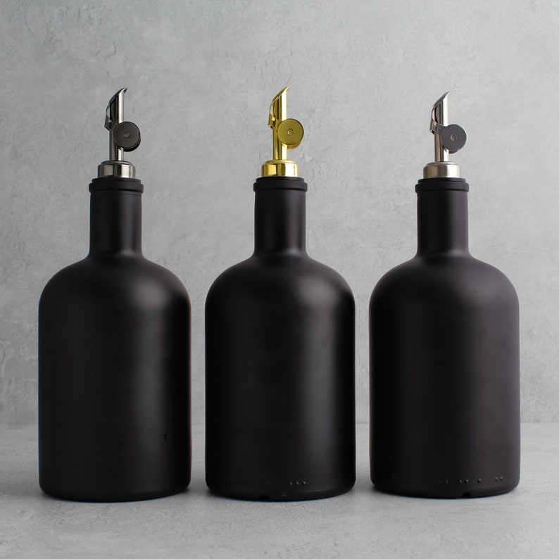 May include: Three black glass bottles with different colored metal tops. The bottles are all the same size and shape, and they are arranged in a row on a gray background.