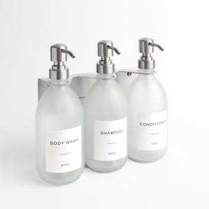 May include: A set of three frosted glass bottles with silver pump dispensers, mounted on a silver metal bracket. Each bottle has a white label with black text: "BODY WASH", "SHAMPOO", and "CONDITIONER". Each bottle holds 500ml.