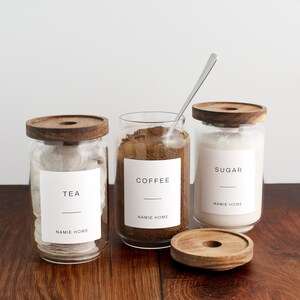 K&ouml;nnte beinhalten: Drei Glasbeh&auml;lter mit Holzdeckeln und Etiketten. Die Beh&auml;lter sind gef&uuml;llt mit Teebeuteln, Kaffeepulver und Zucker. Die Etiketten lauten "Tee", "Kaffee" und "Zucker". Jedes Etikett enth&auml;lt auch den Text "NAMIE HOME".