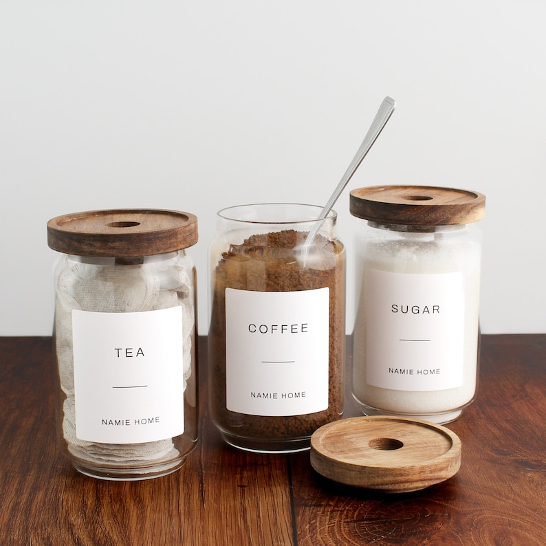 K&ouml;nnte beinhalten: Drei Glasbeh&auml;lter mit Holzdeckeln und Etiketten. Die Beh&auml;lter sind gef&uuml;llt mit Teebeuteln, Kaffeepulver und Zucker. Die Etiketten lauten "Tee", "Kaffee" und "Zucker". Jedes Etikett enth&auml;lt auch den Text "NAMIE HOME".