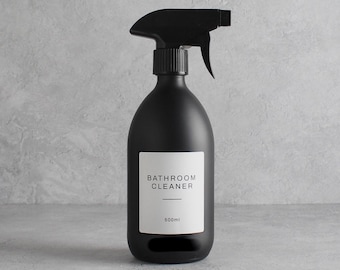 May include: A matte black spray bottle with a black trigger. A white label on the front reads "BATHROOM CLEANER" and "500ml". The bottle has a rounded shape and is designed for cleaning products.