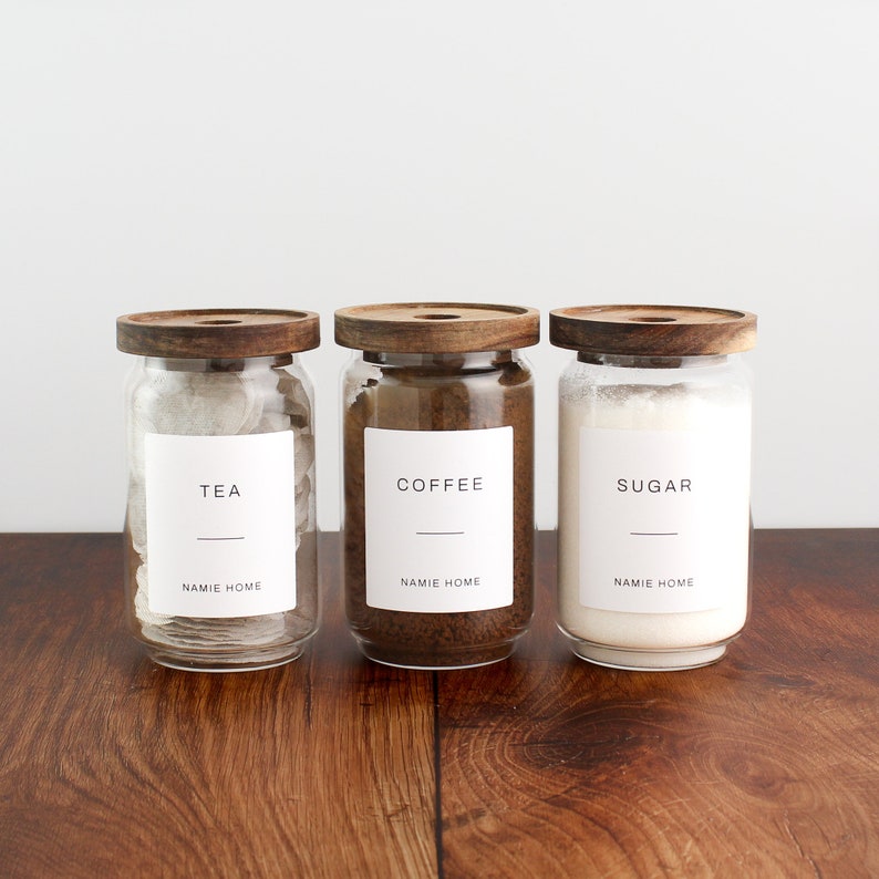 K&ouml;nnte beinhalten: Drei Glasbeh&auml;lter mit Holzdeckeln und Etiketten. Die Beh&auml;lter sind mit Tee, Kaffee und Zucker gef&uuml;llt. Die Etiketten lauten "Tee", "Kaffee" und "Zucker" mit dem Text "NAMIE HOME" unterhalb jeder Etikette.