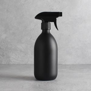 Peut inclure: Un flacon pulvérisateur noir mat avec un mécanisme à gâchette. La bouteille a une forme arrondie et allongée, et la gâchette est noire. Le fond est une surface texturée gris clair.