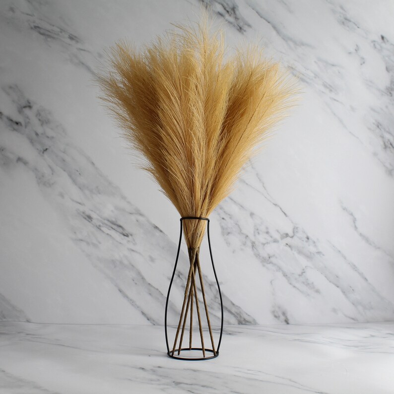 May include: A decorative arrangement of dried pampas grass in a black metal vase. The vase has a slender, open design with a circular base. The pampas grass is a light tan color, and the background is a white marble.