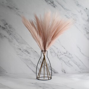 May include: A decorative arrangement featuring a bundle of light pink faux pampas grass in a black metal vase. The vase has a unique wireframe design. The background is a white marble pattern.