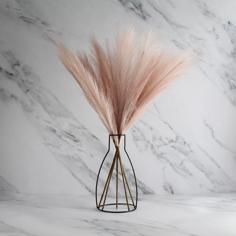 May include: A decorative arrangement featuring a bundle of light pink faux pampas grass in a black metal vase. The vase has a unique wireframe design. The background is a white marble pattern.
