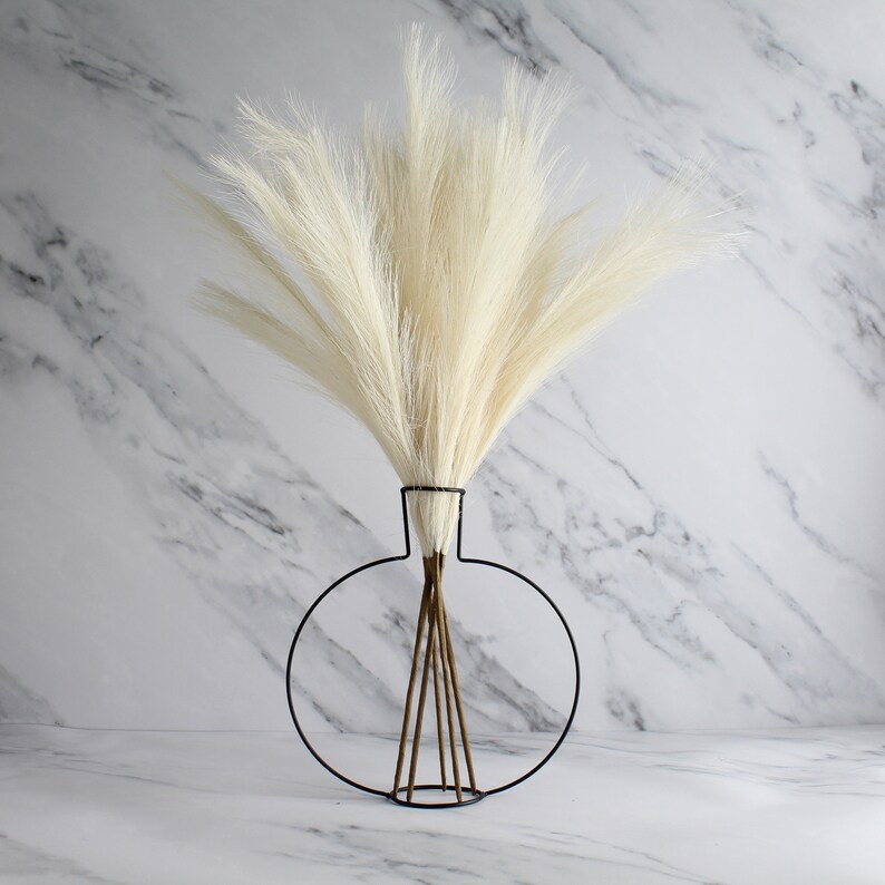 May include: A decorative arrangement featuring tall, cream-colored faux pampas grass in a modern black metal vase. The vase has a circular design with a minimalist aesthetic, and the grass adds a soft, textured element to the composition.