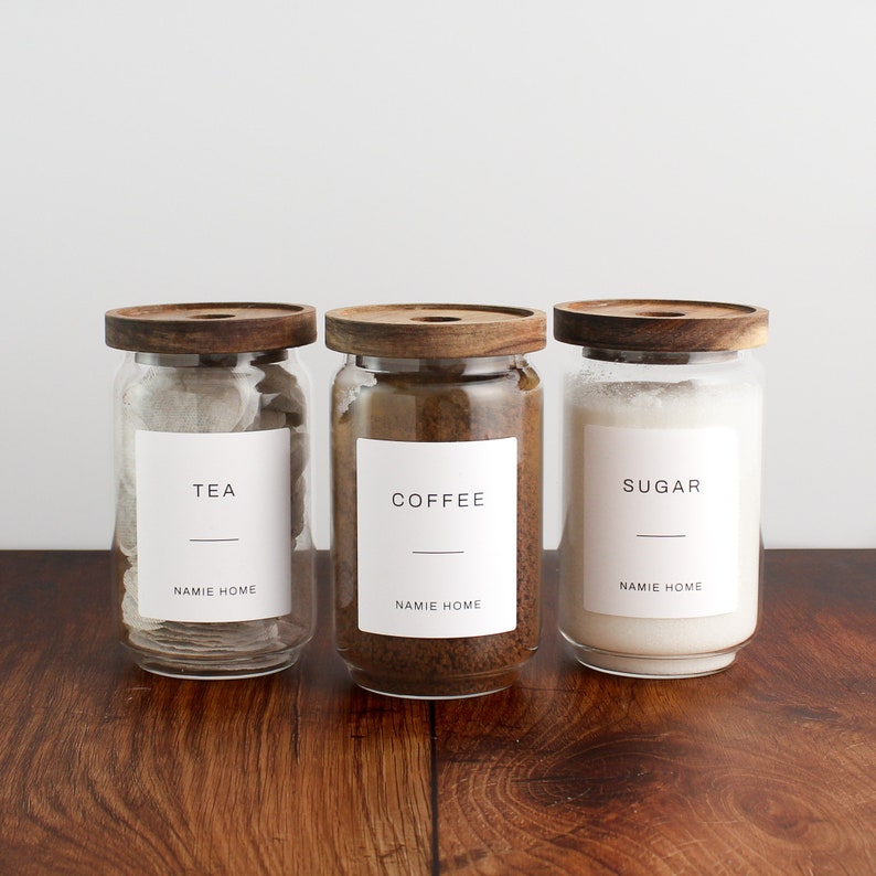 K&ouml;nnte beinhalten: Drei Glasbeh&auml;lter mit Holzdeckeln und Etiketten. Die Etiketten lauten "Tee", "Kaffee" und "Zucker". Jedes Gef&auml;&szlig; tr&auml;gt den Text "NAMIE HOME" auf dem Etikett.