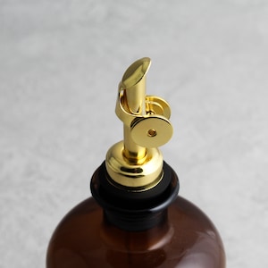 May include: A gold metal bottle stopper with a black rubber ring. The stopper has a spout with a lever that can be used to pour liquids.