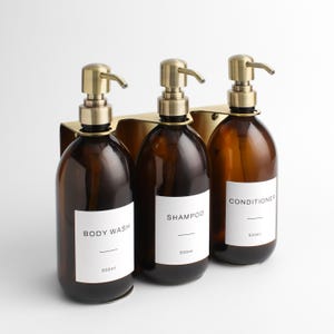 Puede incluir: Tres botellas de vidrio ámbar con dispensadores de bomba color latón, etiquetadas como "Body Wash", "Shampoo" y "Conditioner". Cada botella contiene 500 ml. Las botellas están montadas en un soporte metálico color latón.