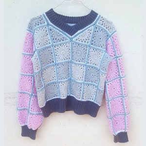 Könnte beinhalten: Ein gehäkelter Pullover mit V-Ausschnitt und langen Ärmeln. Der Pullover ist aus hellblauem und rosafarbenem Garn gefertigt und hat ein quadratisches Muster. Die Ärmel sind einfarbig rosa.