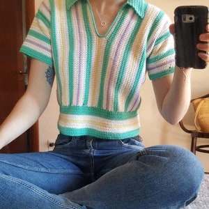 Puede incluir: Un top de crochet a rayas con cuello, mangas cortas y un esquema de color verde, blanco, morado y amarillo. El top es corto y se lleva con jeans azules.