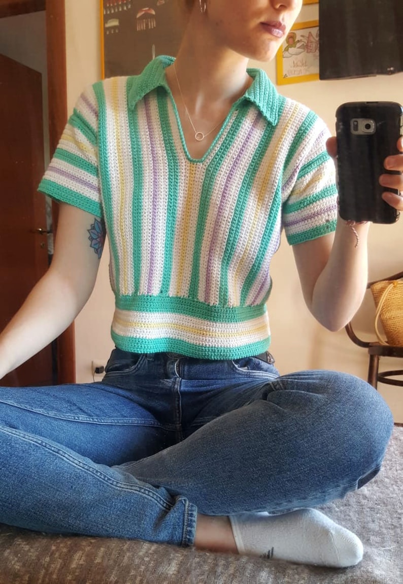 Puede incluir: Un top de crochet a rayas con cuello, mangas cortas y un esquema de color verde, blanco, morado y amarillo. El top es corto y se lleva con jeans azules.