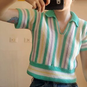 Puede incluir: Un top de crochet a rayas verdes, blancas y moradas con cuello. El top tiene mangas cortas y una longitud recortada.