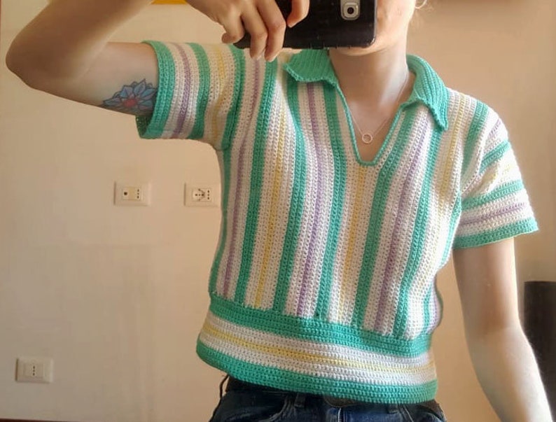 Puede incluir: Un top de crochet a rayas verdes, blancas y moradas con cuello. El top tiene mangas cortas y una longitud recortada.