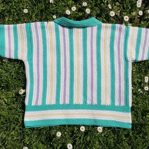 Puede incluir: Un top de crochet a rayas con cuello, mangas cortas y un patr&oacute;n de rayas blancas, amarillas y moradas sobre un fondo azul verdoso.