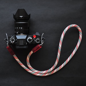 Op de afbeelding: Een zwarte camera met een lens en een rode knop is bevestigd aan een kleurrijk touw camera-riem.