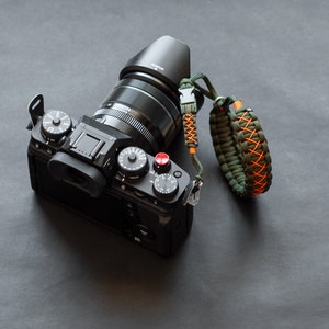 Könnte beinhalten: Eine schwarze Kamera mit einem Objektivdeckel und einem geflochtenen Armband in Grün und Orange.