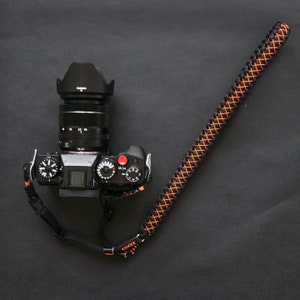 May include: A black camera with a lens and a black and orange camera strap. The camera has a red button and several dials.