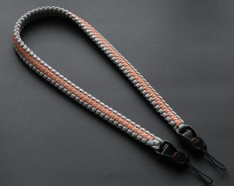 Op de afbeelding: Een gevlochten cameraband in grijs en oranje met zwarte metalen gespen.