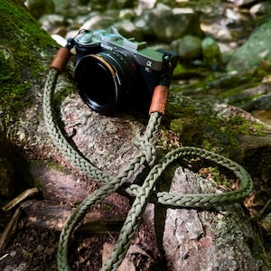 May include: A black Sony Alpha 7C II mirrorless camera with a lens attached, featuring a braided olive green paracord camera strap. The strap includes two brown leather connectors at the attachment points, providing a secure hold for the camera.