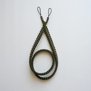 May include: A green and tan braided lanyard with two black loops at the top. The lanyard is made of a thick, durable material and has a loop at the bottom. The design is symmetrical and appears to be handmade.