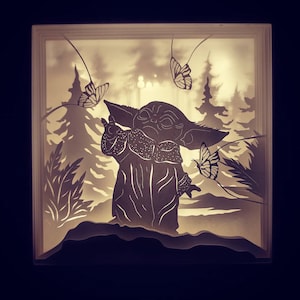 Puede incluir: Una obra de arte en papel en capas que presenta la silueta de un personaje de una popular franquicia de ciencia ficción. La obra de arte representa al personaje en una escena de bosque con mariposas. La pieza está iluminada desde dentro, creando un brillo suave.